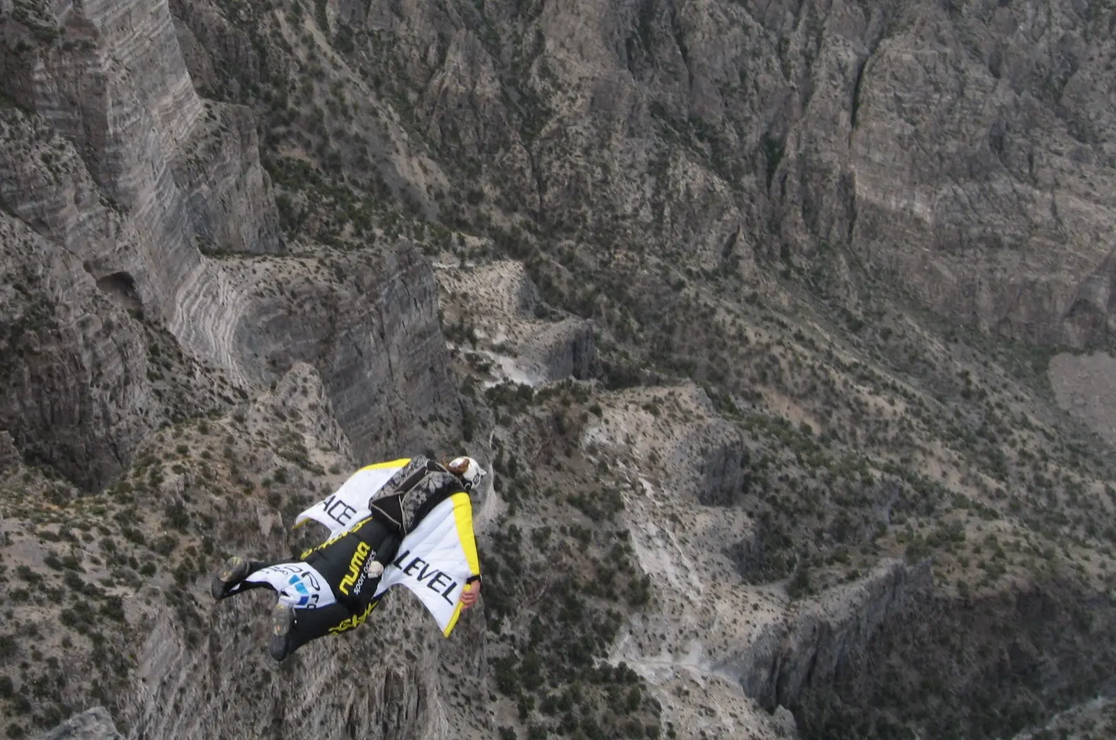 A wingsuit pilot in a yellow and white Face Level wing flying low over a grey canyon