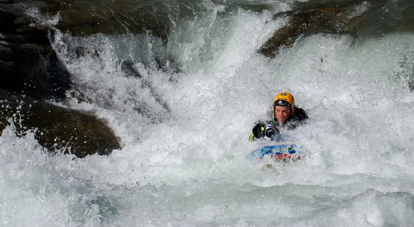 Josh Galt riverboarding headfirst through whitewater, holding a camera and looking straight into the lens