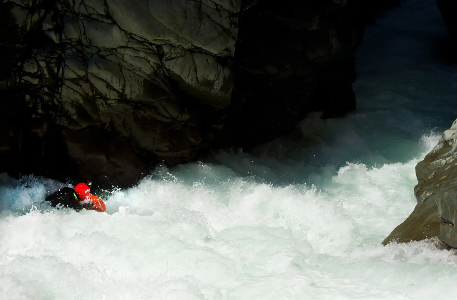 A rider in a red helmet low in the white water of a dark rock chute