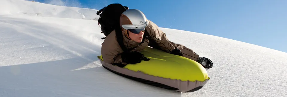 A rider chest-down on a yellow inflatable airboard carving across fresh snow